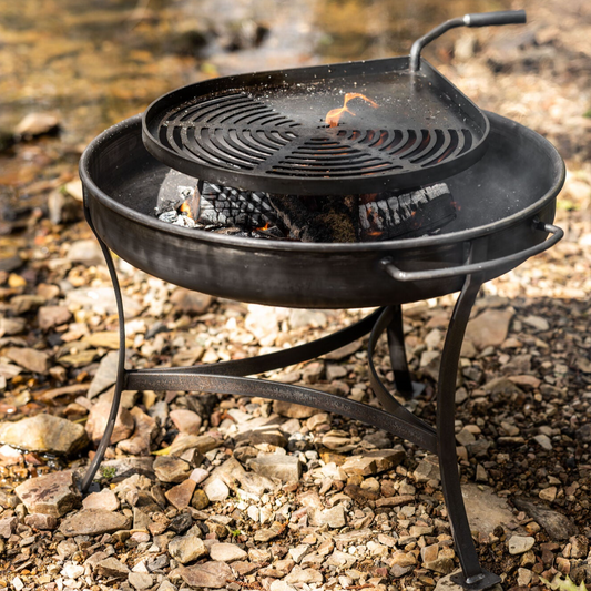 The Moorland fire pit and swing grille set by Dean Forge, featuring a round, black handmade design, is positioned on rocky ground. A lively fire burns beneath the BBQ grill, sending smoke upwards. A river in the background enhances the outdoor, natural setting.