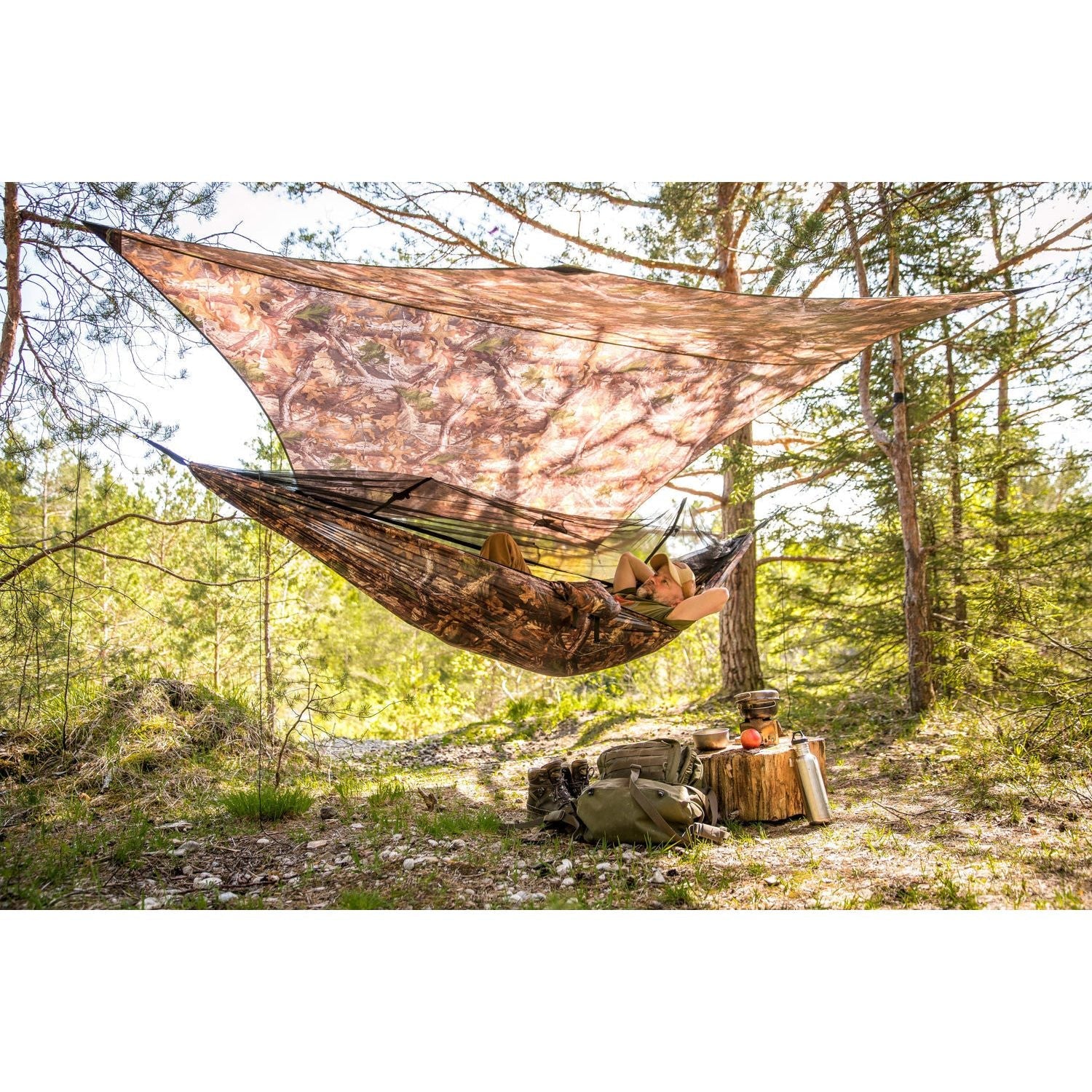 An individual unwinds in the Amazonas Moskito-Traveller Forest hammock, effortlessly strung between two trees within a forest. Above, a tarp offers shade. Close by, on the forest floor, there's a backpack and hatchet next to a log that supports a mug and kettle. Sunlight streams through the trees, adding to the peaceful forest ambiance.