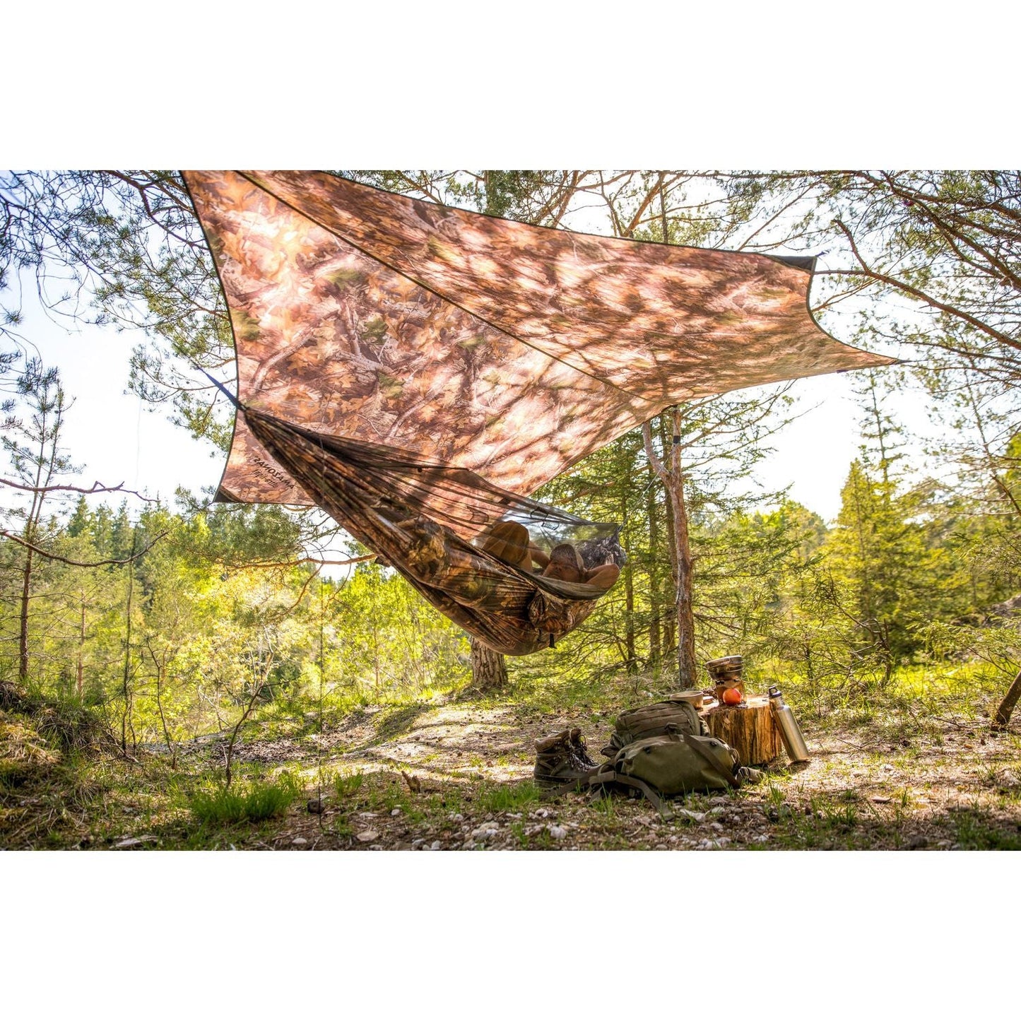 A Moskito-Traveller Forest by Amazonas is set up in the forest, providing a shelter with its ultralight hammock and mosquito net beneath it. The camping scene includes gear like a backpack and food items scattered on the ground amidst lush greenery and trees, with sunlight filtering through the tree canopy.