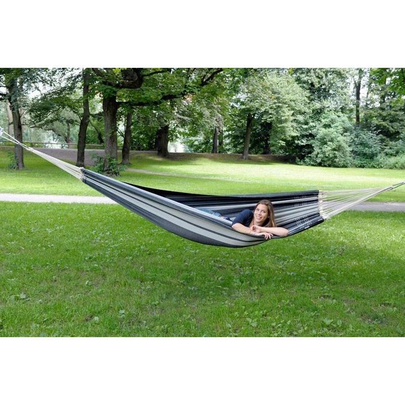 In a lush, green park, someone lounges in an Amazonas Paradiso Silver Hammock crafted from recycled Brazilian cotton, gently swaying between two tall trees. Their smile reveals true contentment as they enjoy this haven for comfort seekers, surrounded by grass and greenery.