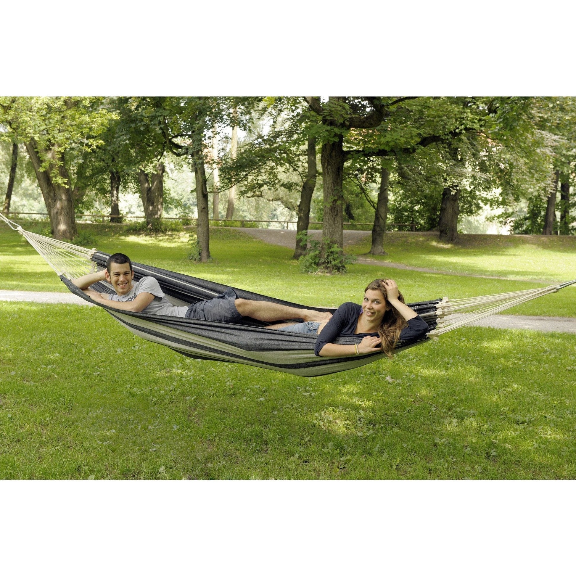 A man and a woman are enjoying a sunny day outdoors, comfortably lounging in an Amazonas Paradiso Silver Hammock, made from recycled Brazilian cotton. The hammock is suspended between trees in a park, where they smile contentedly amidst the lush green grass and trees—a perfect setting for comfort seekers.