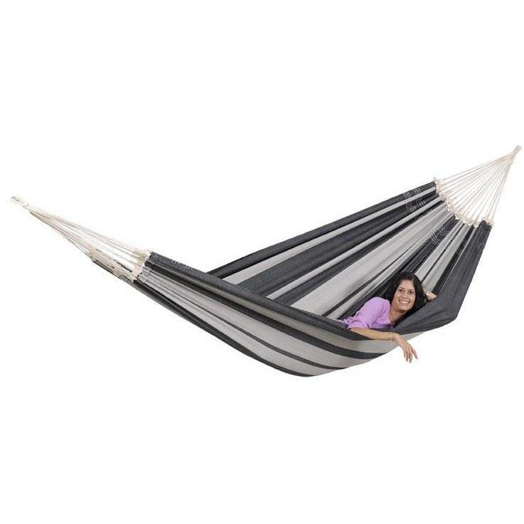 A comfort seeker is unwinding in an Amazonas Paradiso Silver Hammock, crafted from recycled Brazilian cotton. The individual, dressed in a purple top, smiles while savoring the serene moment amidst the hammock's gray-striped fabric—a perfect depiction of leisurely relaxation.