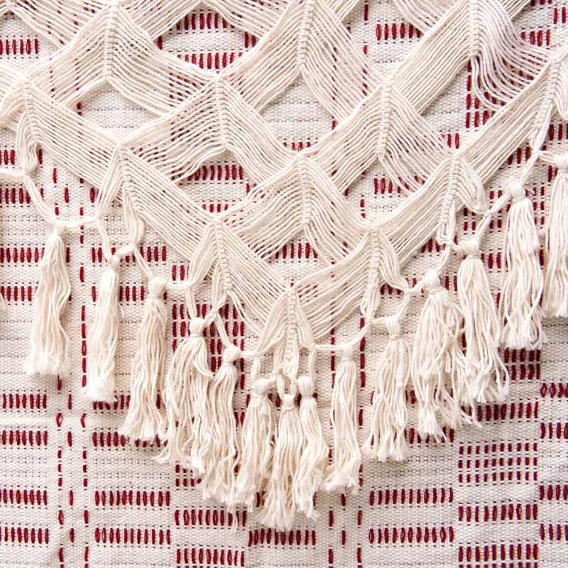 A detailed view of the Rio Bordeaux Hammock from Amazonas showcases its complex cream-colored macramé design, adorned with fringe tassels. The backdrop features a red pattern accentuated by white horizontal and vertical lines, perfect for family use.