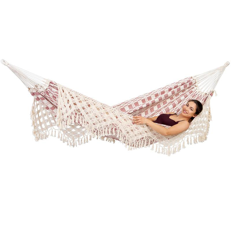 A woman is relaxing in an Amazonas Rio Bordeaux Hammock, featuring beige tones and fringe details. She's smiling and lying comfortably, enveloped by the hammock's intricate patterns against a white background.