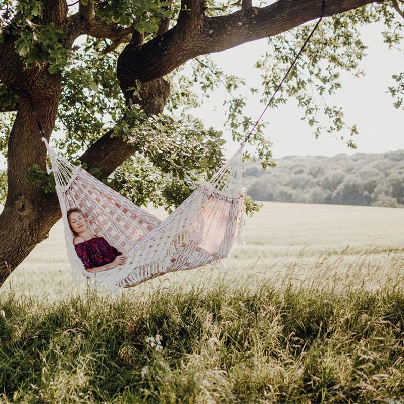 A person relaxes in an Amazonas Rio Bordeaux Hammock strung between the branches of a large tree. They are surrounded by a grassy field and open landscape, perfect for family use. The scene is peaceful, with dappled sunlight filtering through the leaves.