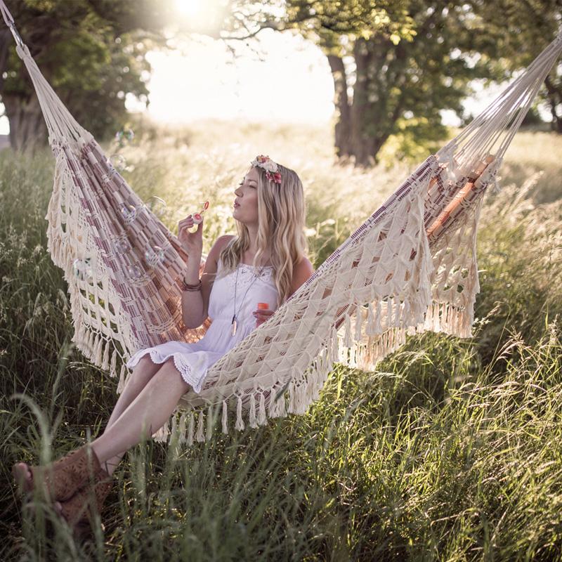 A woman wearing a white dress enjoys relaxing in an Amazonas Rio Bordeaux Hammock, surrounded by lush greenery and soft sunlight filtering through the trees. She blows bubbles in the sunlit grassy field, adorned with a flower headband and ankle boots.