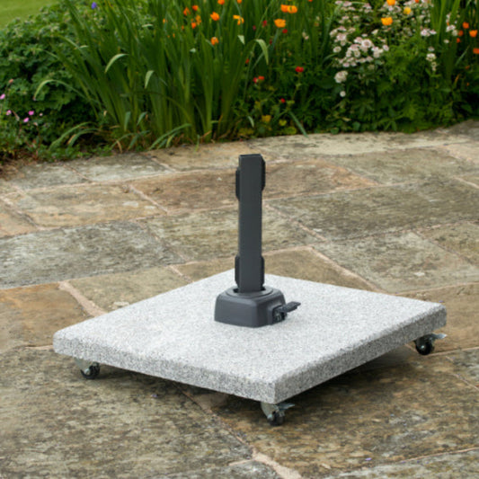 The Salerno Hammered Grey Granite 90KG Wheeled Parasol Base by Pacific Lifestyle is elegantly positioned on the patio, composed of square gray stone and featuring a central black vertical fixture. Surrounded by lush green plants and vibrant flowers, it comes equipped with an adjustable turn lock to perfectly secure your shade.