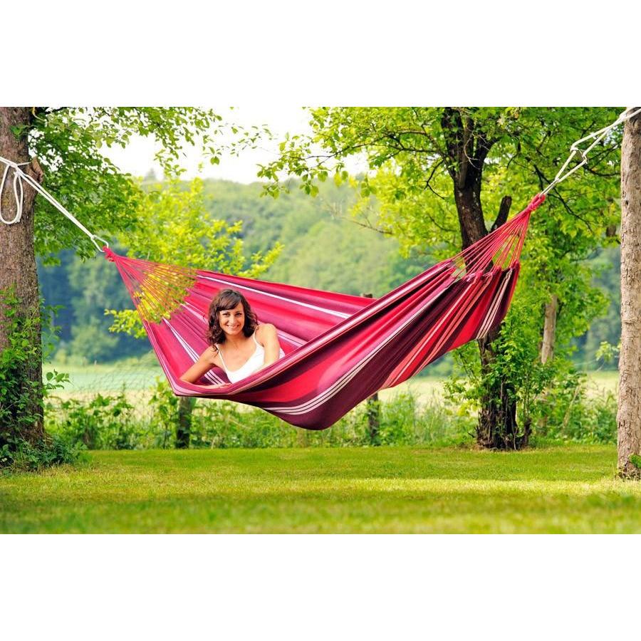 A person enjoys a peaceful moment in an eye-catching pink and red Salsa Fuego Hammock by Amazonas, suspended between two trees in a verdant green park. Crafted from durable, weatherproof EllTex material, the hammock withstands the elements effortlessly. The backdrop showcases expansive grass and lush foliage, enhancing the bright and tranquil ambiance.