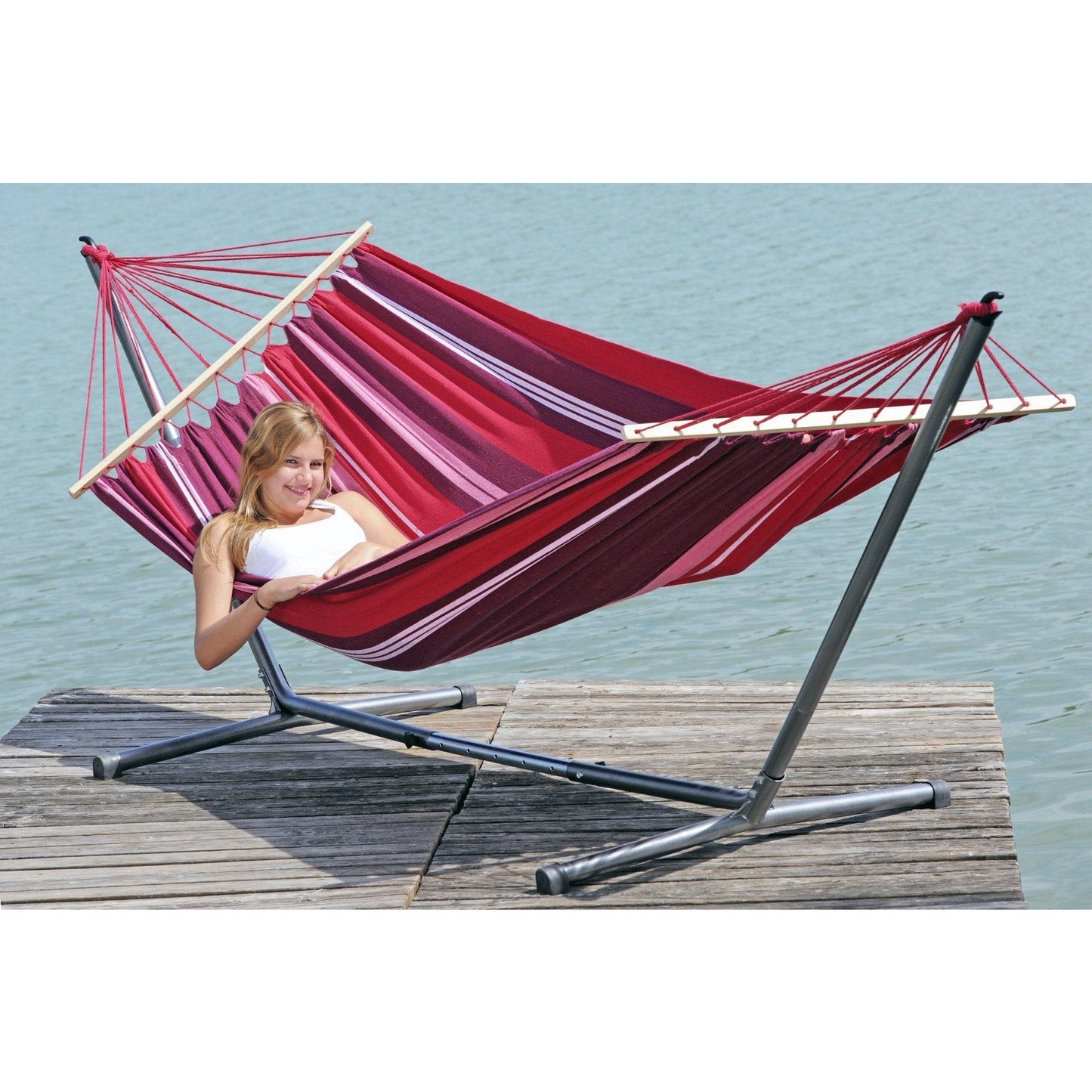 A person relaxes in the Amazonas Samba Fuego Hammock, featuring weather-resistant red and purple stripes, supported by a metal stand on a wooden dock by a calm body of water.