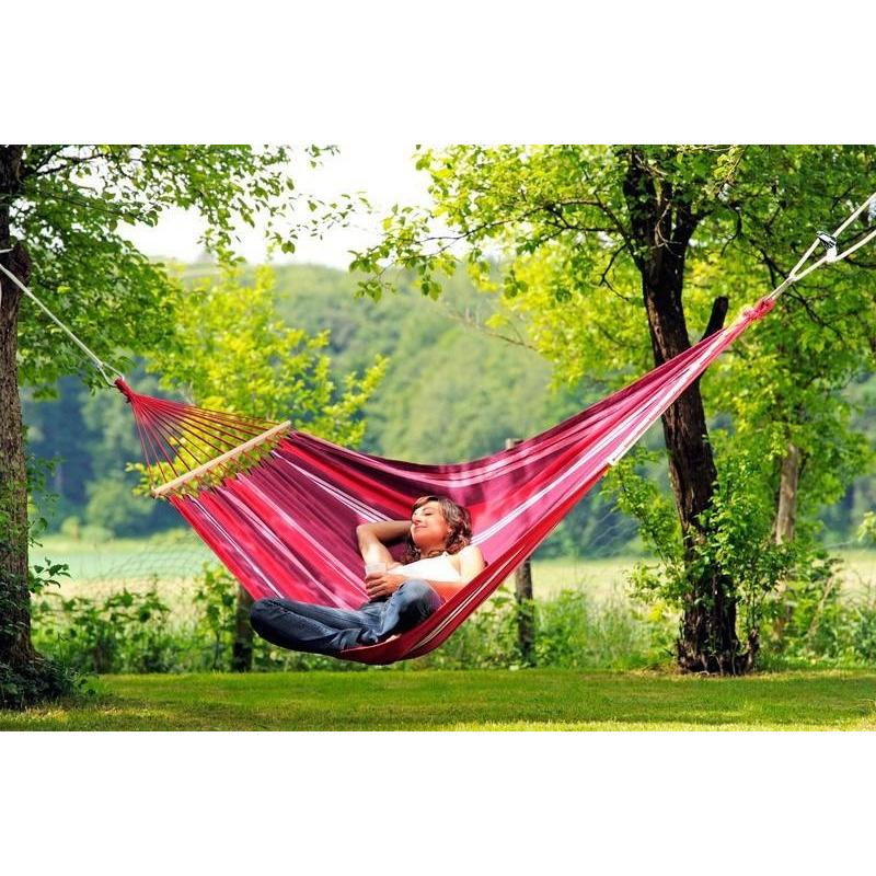 Nestled in a cozy Samba Fuego Hammock by Amazonas, an individual is unwinding between two trees. The hammock's vibrant red and pink stripes, made from durable EllTex fibres, blend seamlessly with the lush green foliage and grass. They savor the forest view under a sunny sky, assured of their weather-resistant sanctuary's ability to endure the elements.