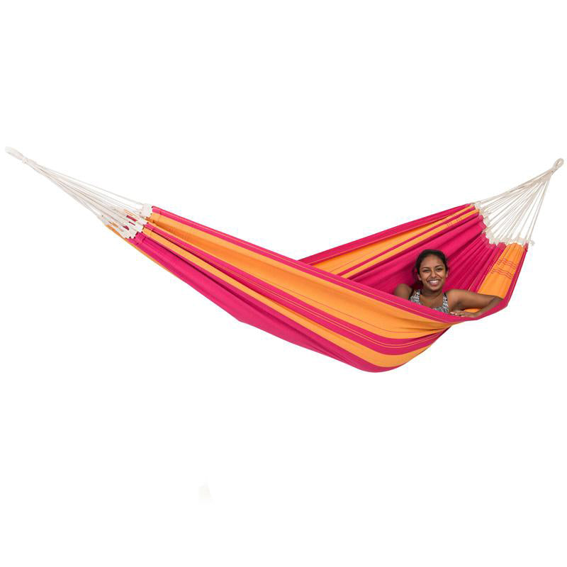 A person smiling while lounging in the vibrant and colorful Santana Hammock Pink by Amazonas, featuring striking stripes of orange, pink, and yellow against a white backdrop. Embracing the cheerful comfort of this exquisite hammock.