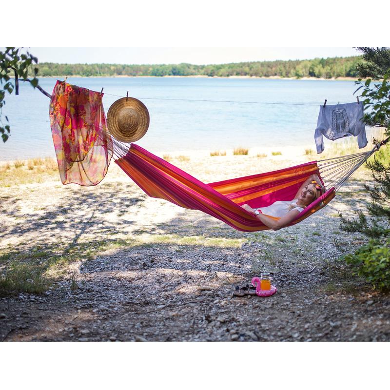 A person relaxes in a vibrant Santana Hammock Pink by Amazonas next to a lake. Clothes and a straw hat hang on a line above, contributing to the colorful comfort. Trees frame the tranquil waterside scene, with a pair of shoes resting on the ground nearby.