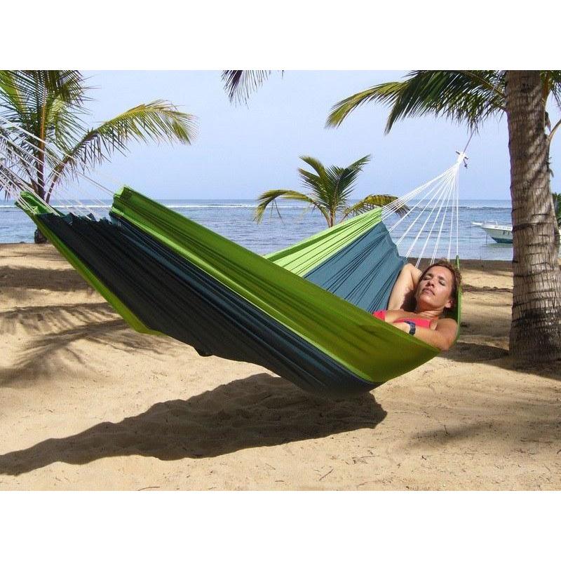 A person relaxes in an Amazonas Silk Traveller Forest Hammock on a sandy beach. Palm trees and a calm sea are in the background under a clear sky, creating a tranquil tropical setting perfect for any outdoor enthusiast.