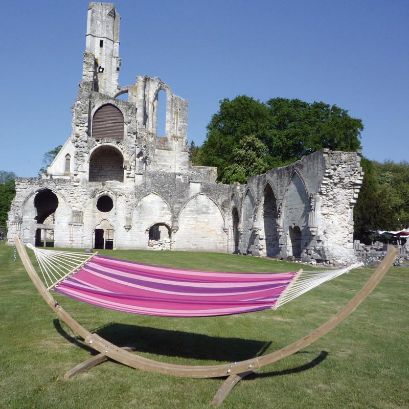 The **Star Candy Hammock Set** by **Amazonas**, featuring a vibrant pink and purple striped design, is crafted from durable Elltex fibres and suspended on a spruce wood stand resting on a lush green lawn. In the background, ancient stone ruins with arches and a partially collapsed tower grace the scene under a clear blue sky.