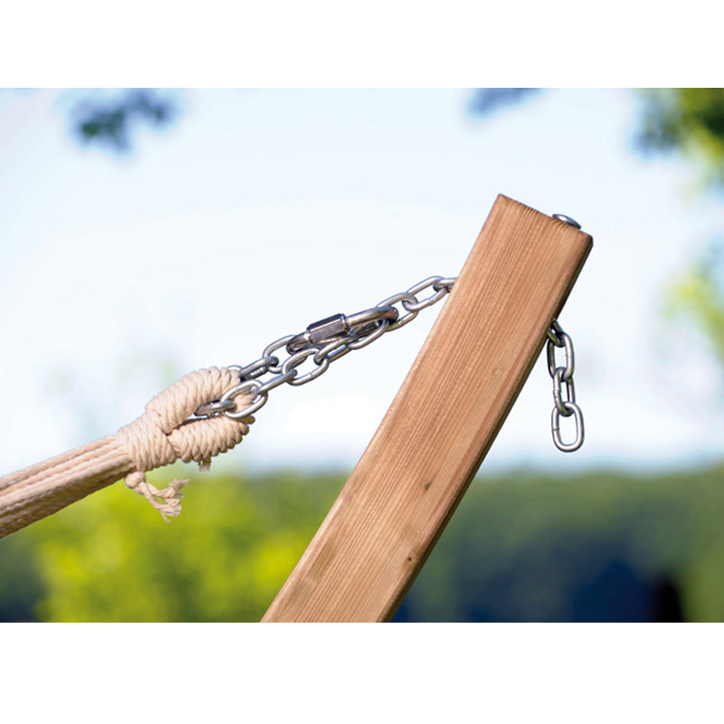 A close-up of the Star Candy Hammock Set by Amazonas showcases a spruce wood stand, complete with a metal chain and durable Elltex fibers holding the hammock securely. The background highlights a garden adorned with lush greenery against a bright, clear sky.