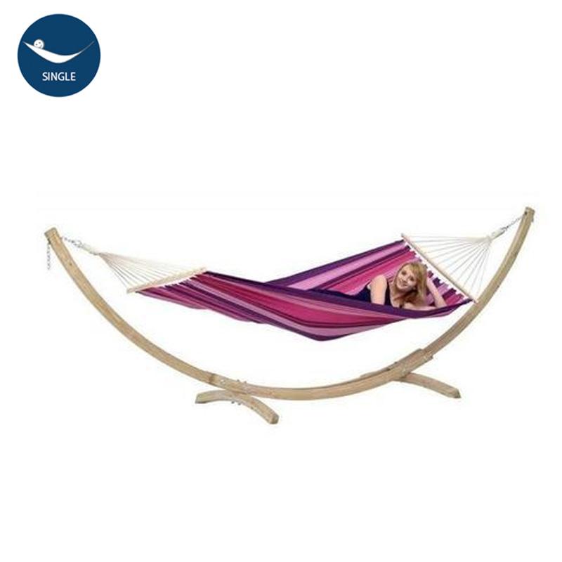 In her garden, a woman lounges comfortably in the Star Candy Hammock Set by Amazonas, showcasing vibrant stripes of pink, purple, and white. The hammock is gracefully supported by a durable spruce wood stand and features an eye-catching design crafted from Elltex fibers. A Single logo is prominently displayed in the top left corner.