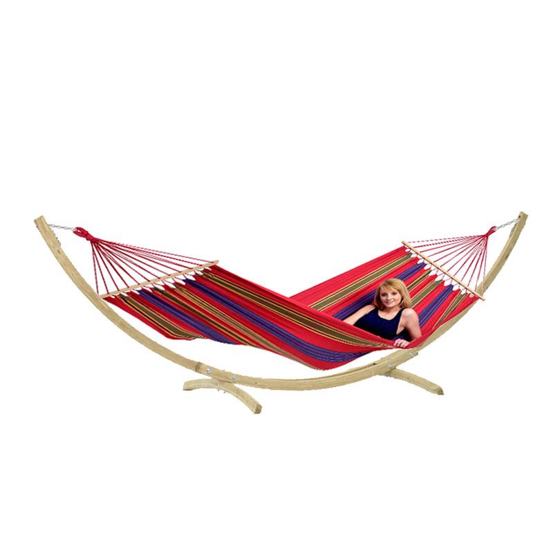 A woman lounges on the Star Hammock Set from Amazonas, suspended on a wooden stand. This vibrant garden addition features weatherproof stripes in red, blue, green, and yellow. Reclining comfortably with a smile, she savors the tranquility of the moment.