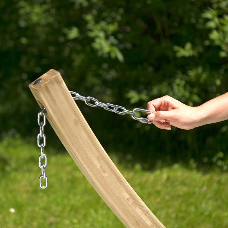 A hand holds a metal chain connected to the elegantly curved wooden beam of the Amazonas Star Hammock Set, forming a stunning garden feature against a backdrop of lush green grass and softly blurred foliage. This weatherproof product offers both durability and charm for outdoor spaces.