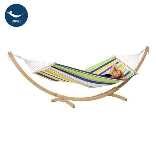 A person relaxes in the Star Kolibri Hammock Set by Amazonas, featuring a striped design crafted from durable EllTex fibers on a sturdy spruce wood stand. The hammock displays vibrant green, blue, yellow, and white stripes with a single icon in the top left corner.