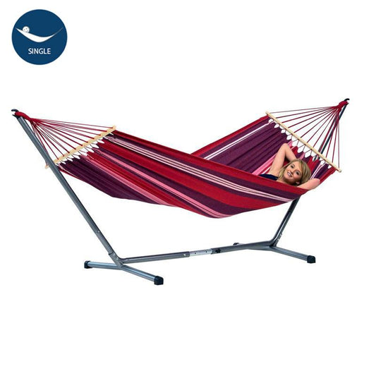 An individual lounging in the Amazonas Summer Hammock Set, featuring a red-striped design and a portable metal stand. Made from weather-resistant EllTex fibers, this hammock is designed for single use.