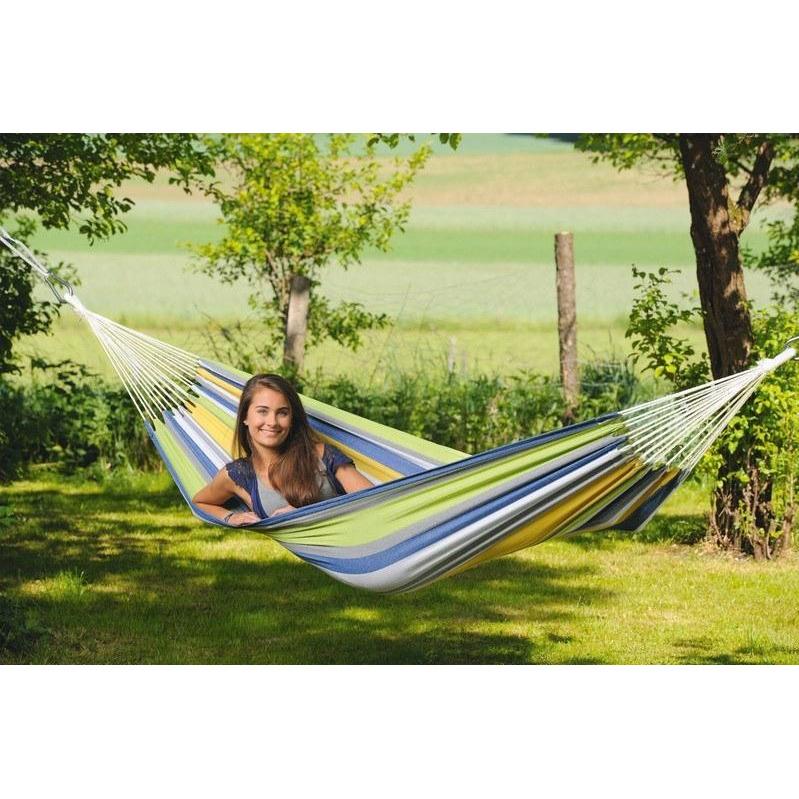 A person relaxes in an Amazonas Tahiti Kolibri Hammock, its colorful strands forming a cocoon between two trees in a verdant meadow, set against a picturesque backdrop of grass and trees, offering both comfort and weather resistance.