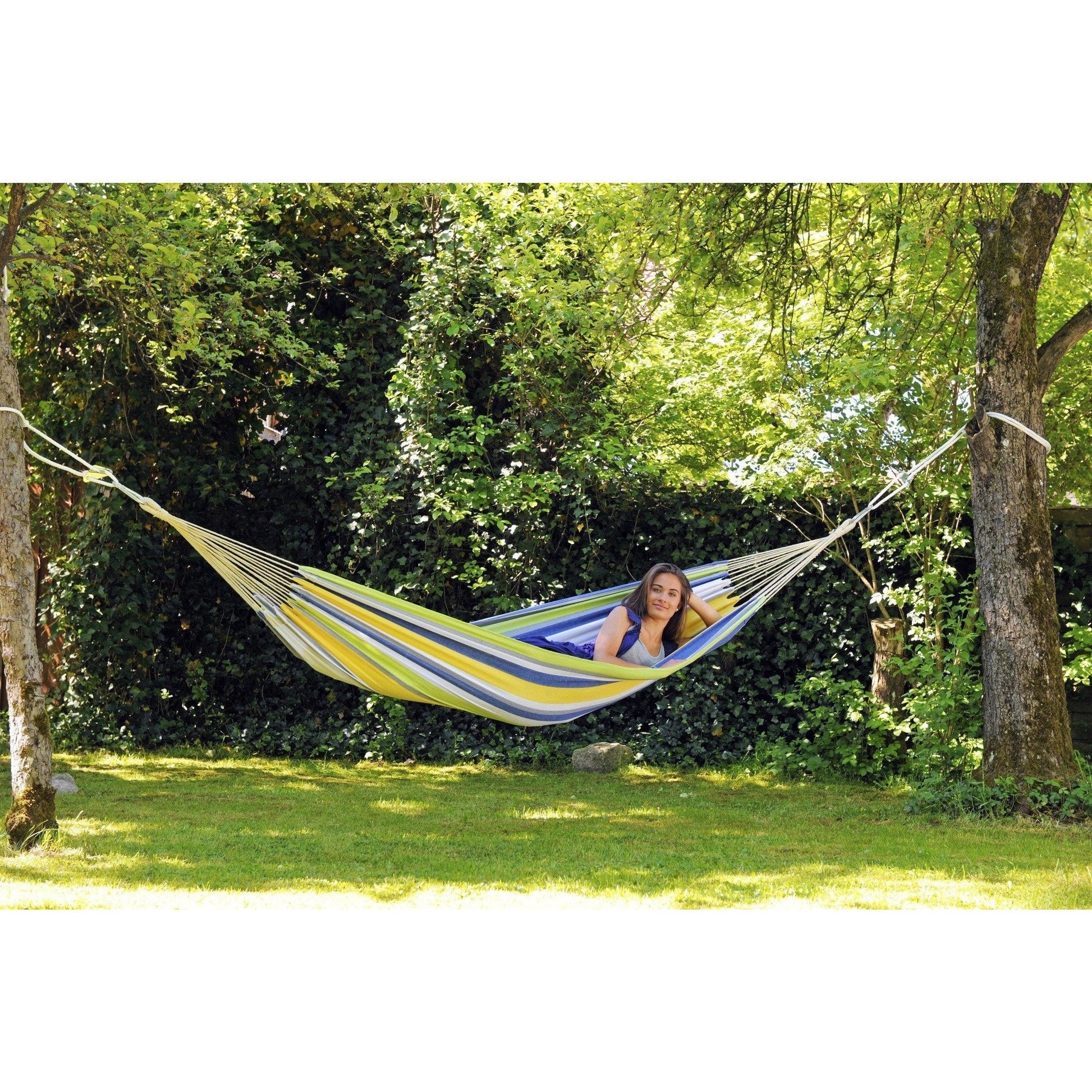 A person relaxes in an Amazonas Tahiti Kolibri Hammock, which is tied between two trees in a lush, green garden. The weather-resistant fibers of the hammock sway gently as sunlight filters through the leaves, creating a peaceful and idyllic outdoor setting.