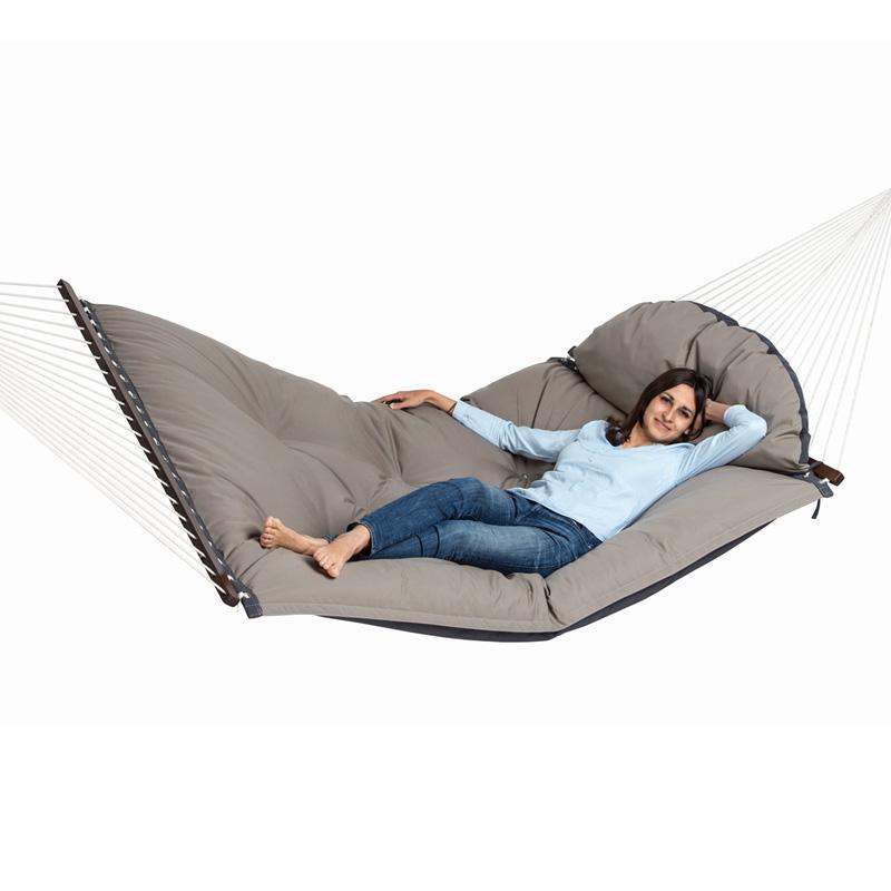 A woman in a light blue sweater and jeans relaxes on "The Fat Hammock - Reversible" by Amazonas, a large, cushioned gray hammock quilt. She is smiling with one arm resting behind her head as the hammock is suspended by ropes on each side, providing an inviting retreat for luxury seekers.