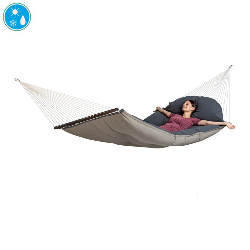 A person is comfortably lounging in The Fat Hammock - Reversible by Amazonas, featuring a beige quilt with a dark cushion and suspended by white ropes. Dressed in a purple shirt, the individual looks perfectly content while resting outdoors. An icon indicates its water-repellent qualities, making this luxurious hammock ideal for any weather condition.