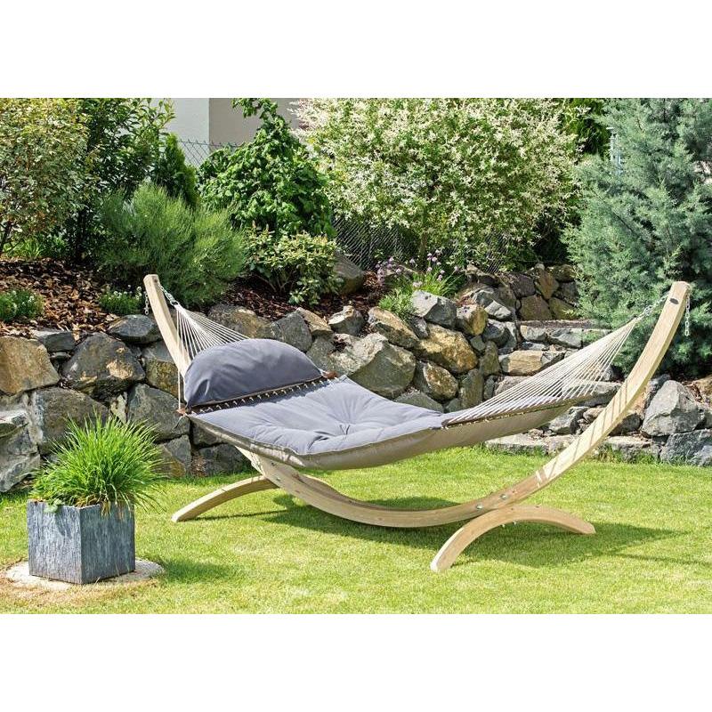 The Fat Hammock - Reversible by Amazonas, featuring a gray, water-repellent cushioned quilt, is elegantly displayed on a wooden stand on a green lawn. In the background, a stone wall adorned with lush shrubs and plants adds to the setting's charm. To the left, a small square stone planter filled with grass beckons luxury seekers to unwind in style.