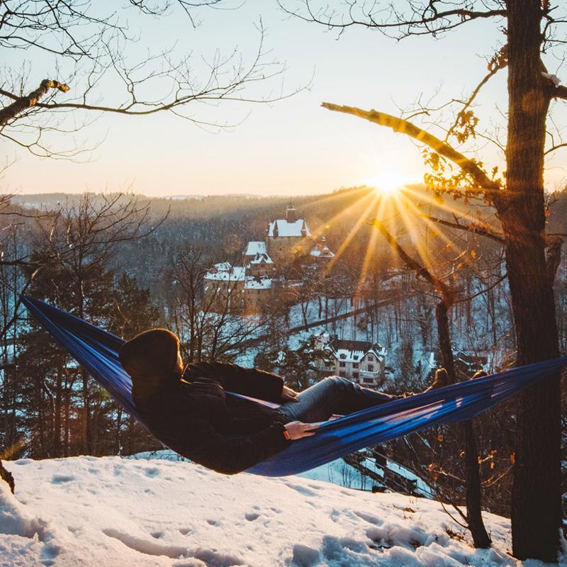 The Travel Hammock Set Blue by Amazonas gently cradles a person between two trees on a snowy hillside. As the sun sets, casting golden light over the forest and distant buildings, this peaceful scene is perfect for outdoor enthusiasts seeking serenity in nature.