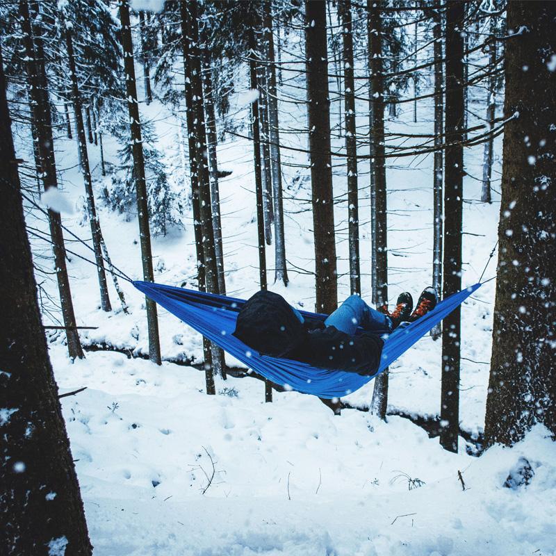 Outdoor enthusiasts find a true retreat by settling into the Travel Hammock Set Blue by Amazonas, made of durable ripstop nylon. Surrounded by snow-covered trees and gentle falling snowflakes, they enjoy a peaceful moment while warmly bundled in a black jacket and boots, embraced by the tranquility of the forest.