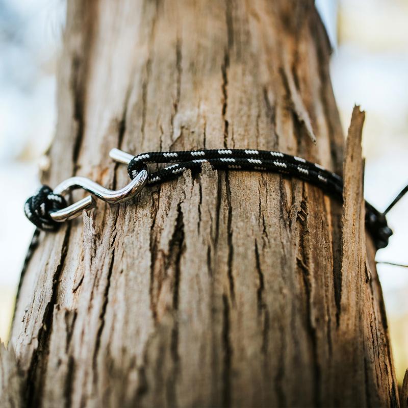 A close-up of a black and white braided rope, crafted from ripstop nylon, fastened with a hook around the rough, textured surface of a tree trunk. The rope's design contrasts strikingly with the tree's natural ruggedness—an ideal arrangement for outdoor enthusiasts using their Amazonas Travel Hammock Set Blue.