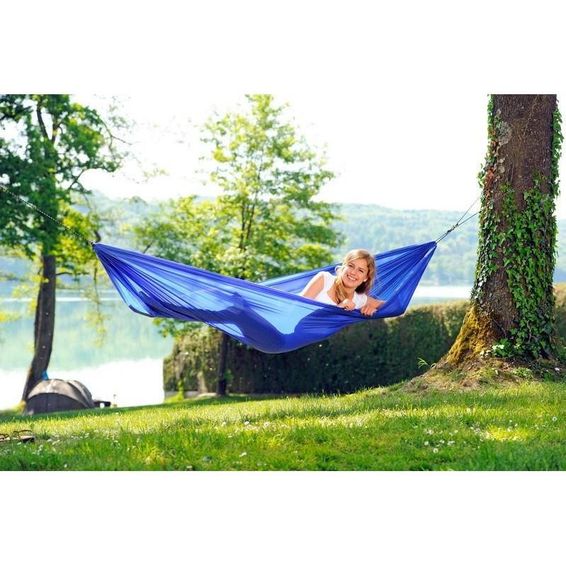 An Amazonas Travel Hammock Set Blue, crafted from resilient ripstop nylon, gracefully sways between two trees, supporting a person above the verdant grass. Sunlight filters through the leaves, creating a pattern of dappled light over the tranquil setting of trees and a shimmering lake—a perfect retreat for outdoor enthusiasts in search of tranquility.